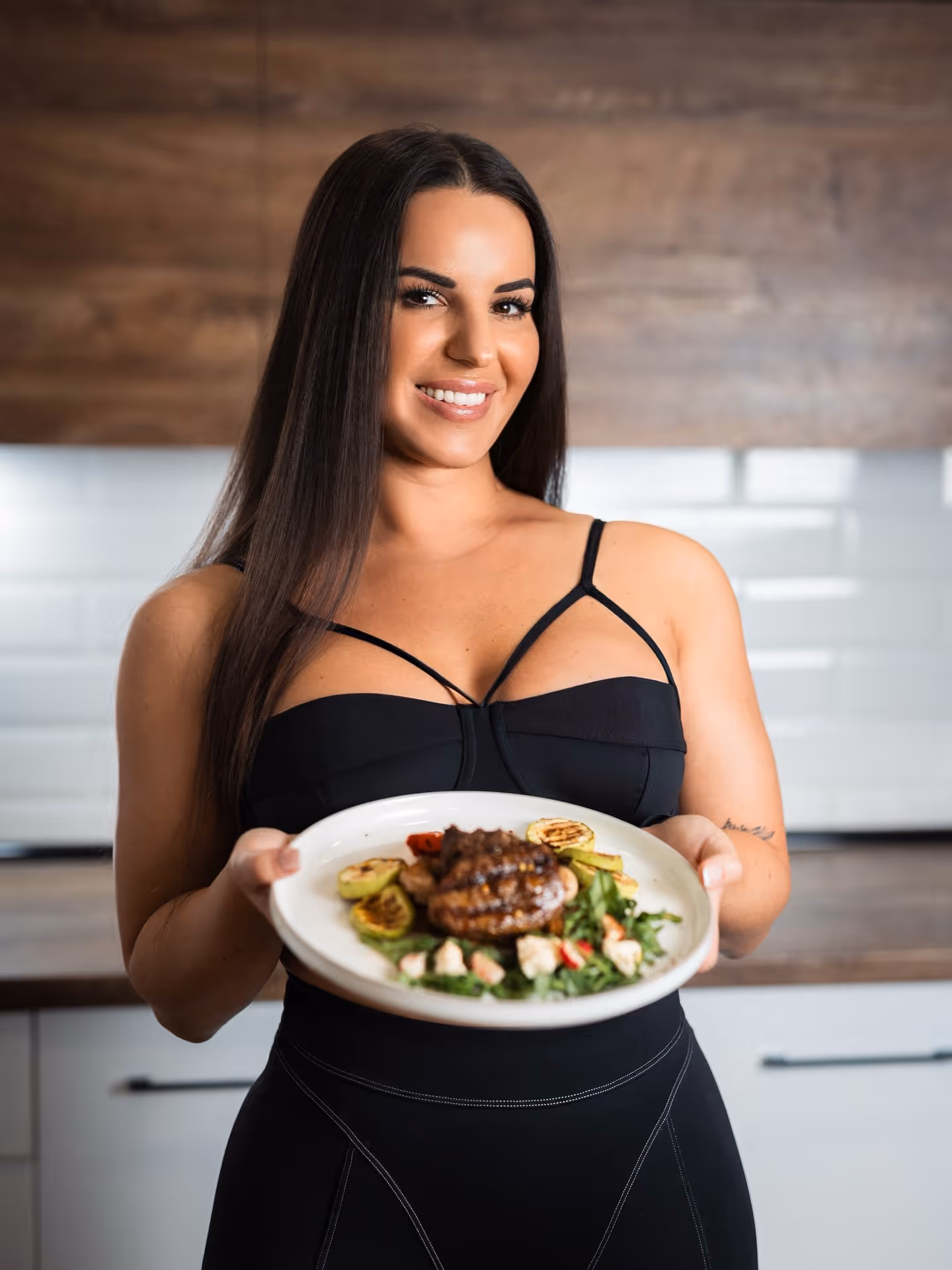 Smiling woman in black sportswear holding a plate with grilled vegetables and meat in a kitchen.